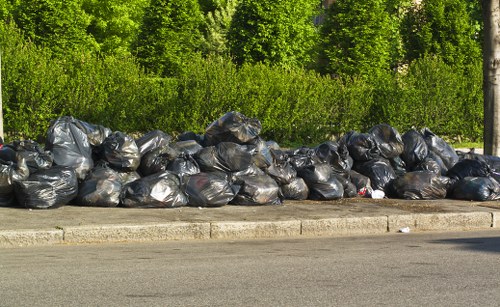 A row of black rubbish bags filled with waste material, stacked along the edge of a paved roadside area, with a curb separating them from the asphalt. Behind the bags, there is a dense green hedge and tall trees, indicating the setting is outdoors. The rubbish bags are made of plastic, appear tightly packed, and are situated in a straight line on the pavement, suggesting a rubbish removal or collection point. The natural lighting highlights the sheen on the plastic bags and the vibrant greenery in the background. This scene aligns with the services offered by Shoreditch House Clearance for rubbish disposal or clearance in the local area, with the image capturing an environment that might be typical of waste awaiting collection or disposal in East London.