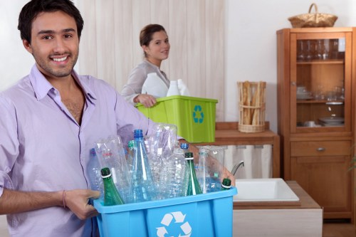 A young man with short dark hair and a friendly expression is standing in a well-lit kitchen, holding a blue recycling bin filled with clear plastic bottles, some with green and blue labels visible. Behind him, a young woman with brown hair tied back is placing a green recycling bin on a countertop; she is dressed casually, and the bin has a white recycling symbol on the front. The kitchen features light-colored wood cabinets, a wooden display cupboard on the right, and a white sink with a silver faucet. The background wall is covered with light-colored vertical paneling, and natural light illuminates the space. The scene depicts the process of sorting recyclables, relevant to rubbish removal services offered by Shoreditch House Clearance, especially in urban areas like Shoreditch or nearby districts, emphasizing environmentally conscious waste management.