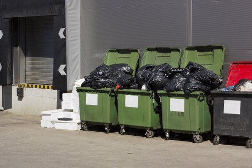 A row of four green wheelie bins with open lids is positioned on a concrete driveway or pavement area, filled with black rubbish bags and miscellaneous waste materials. The bins are aligned side by side, with some bags protruding from the tops. To the right, a smaller black waste bin with a white label is also visible, and behind the bins, there is a grey industrial shutter door with a yellow-and-black striped safety marking at its base. The background includes a modern building with grey paneling, suggesting a commercial or residential property in Shoreditch. The scene is illuminated by natural daylight, emphasizing the textures of the plastic bins and waste bags, conveying a typical rubbish collection or clearance scenario managed by Shoreditch House Clearance as part of their local rubbish removal services.