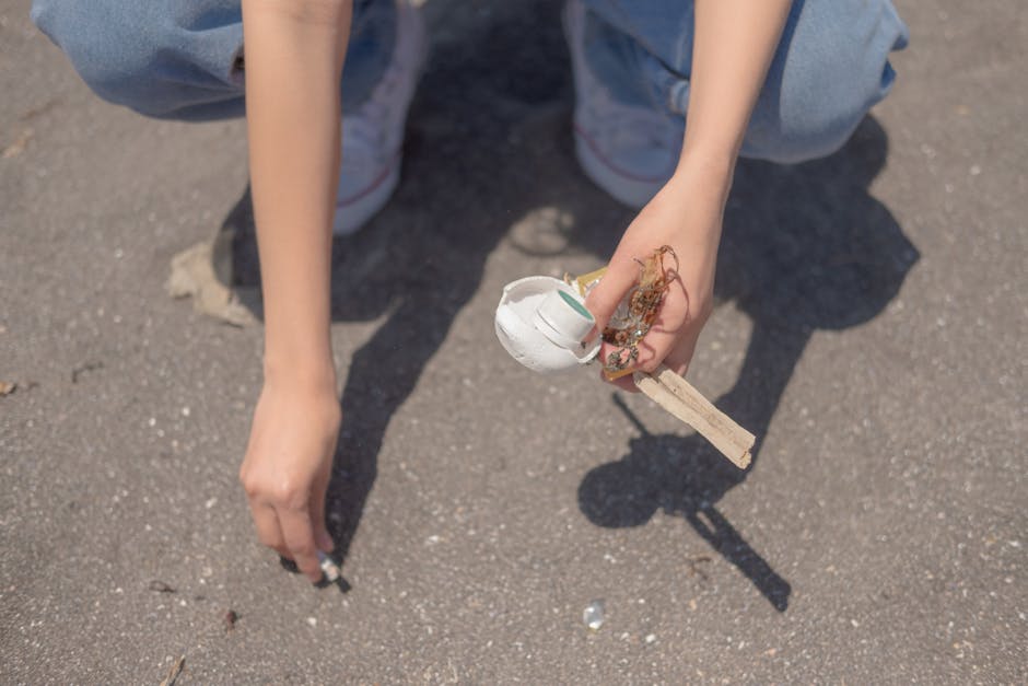 A person, wearing light blue denim jeans and white sneakers, is crouching on an outdoor paved surface, holding a broken ceramic mug with a white glazed finish and a green decorative detail, along with