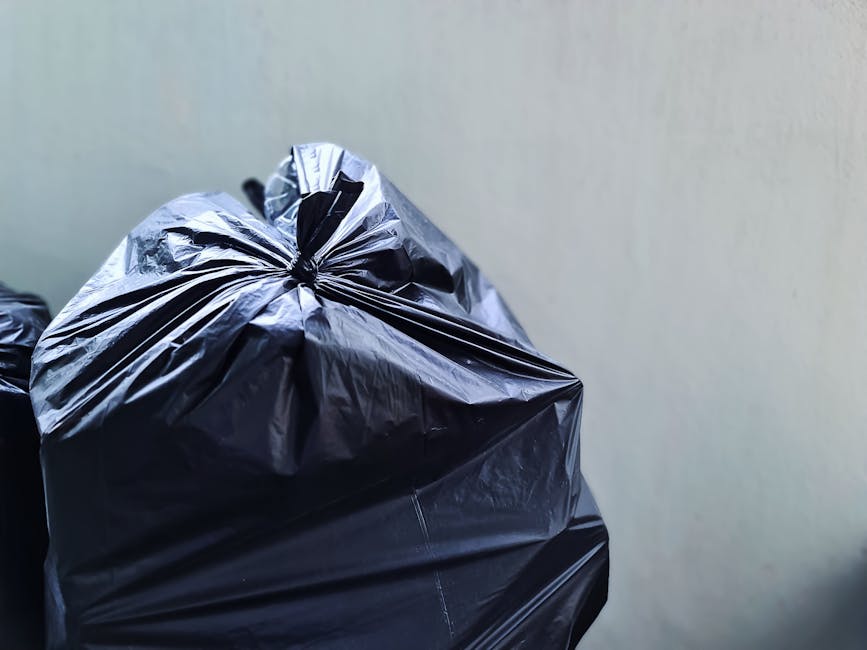Close-up image of several large black plastic rubbish bags filled with waste, tightly knotted at the top, with a smooth, shiny texture reflecting light. The bags are positioned on a flat, grey surface
